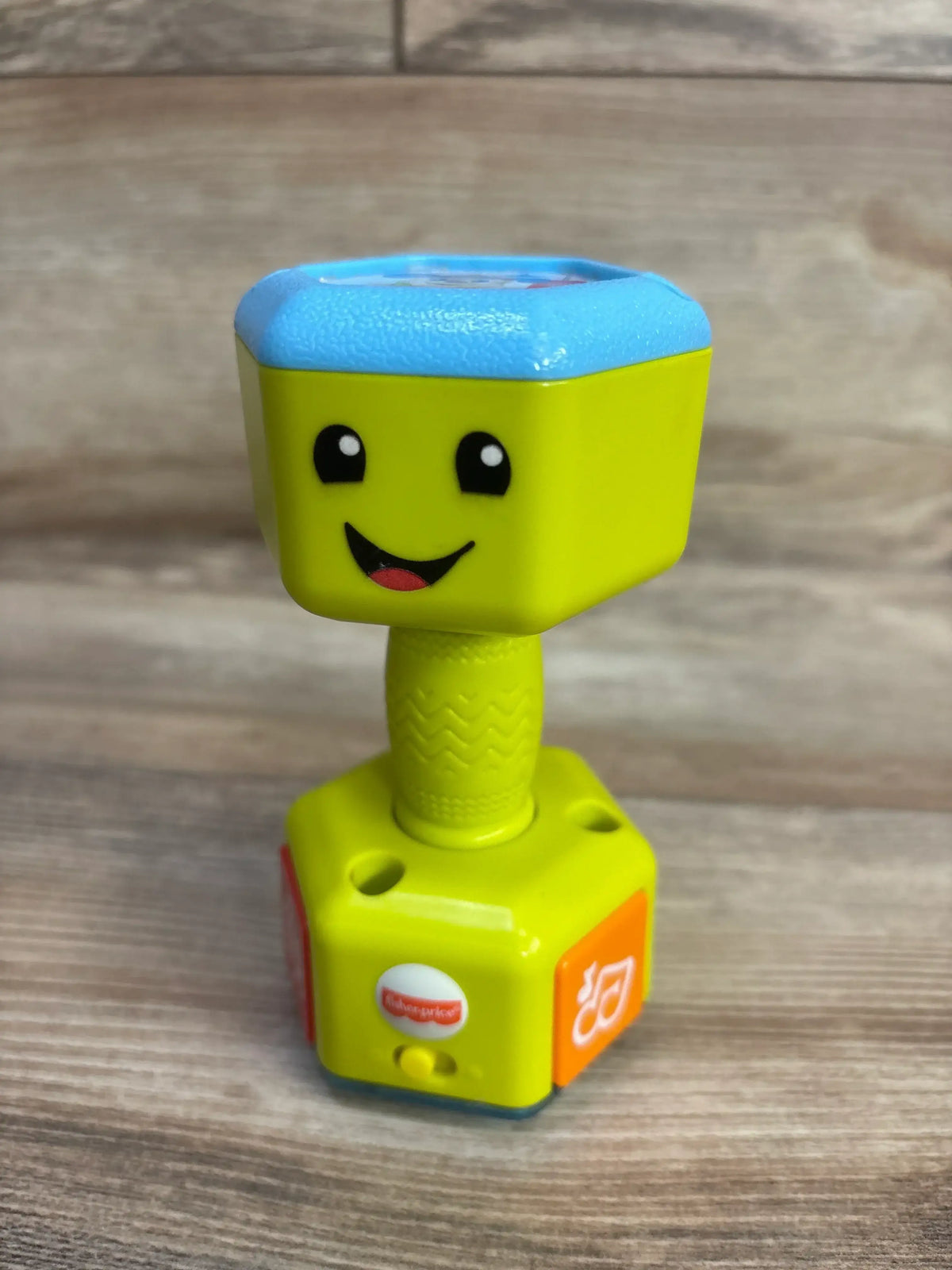 Front view of Fisher-Price Countin Reps Dumbbell Rattle green showcasing smiley face with blue top on wood plank floor