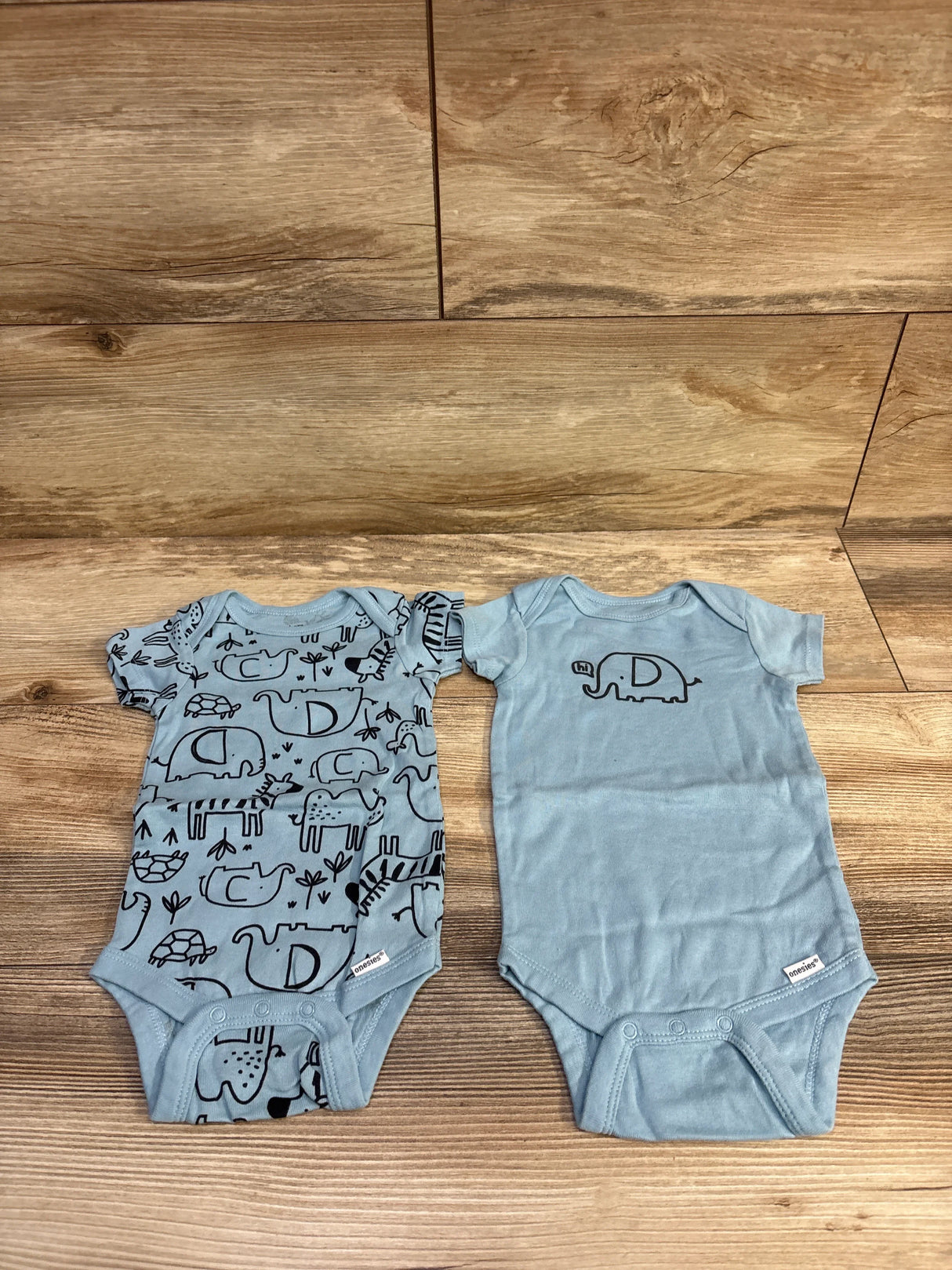 Flat lay of Gerber 2-pack blue baby bodysuits, one with black animal print and one solid with small elephant on wood floor