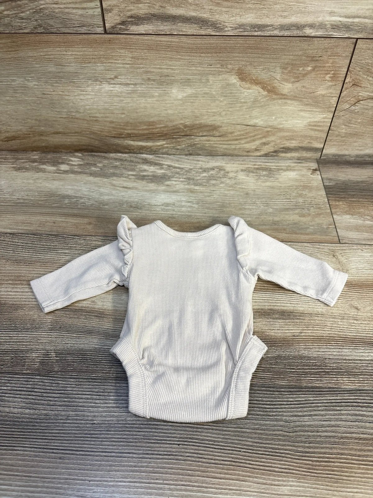 Flat lay of beige preemie ribbed long-sleeve bodysuit showing ruffle shoulder detail on a wood-plank background