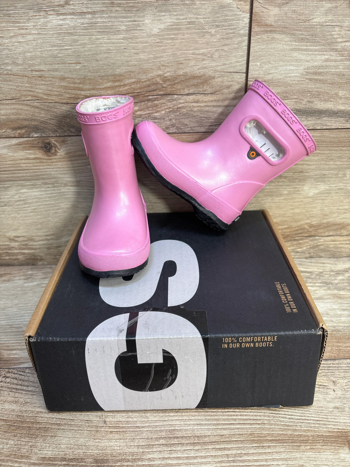 Pink Bogs kids rain boots with handles and cozy lining on a black shoe box, wood background