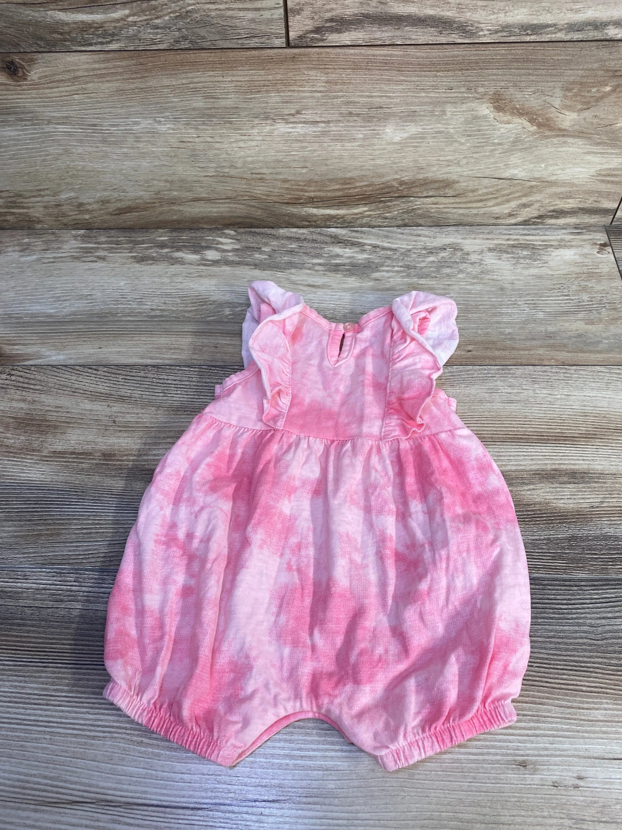 Front flat lay of pink tie-dye shortie romper with ruffle shoulders, gathered waist and elastic leg openings on wood