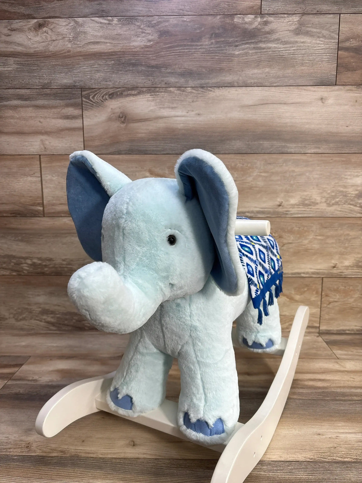Front view of light blue plush elephant rocker with large ears, blue patterned saddle and white wooden base on wood floor