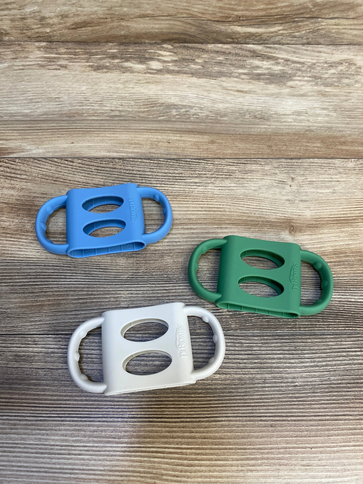 Top-down view of three Dr. Brown’s silicone narrow bottle handles in blue, green, and white showing dual hand grips on a wood surface