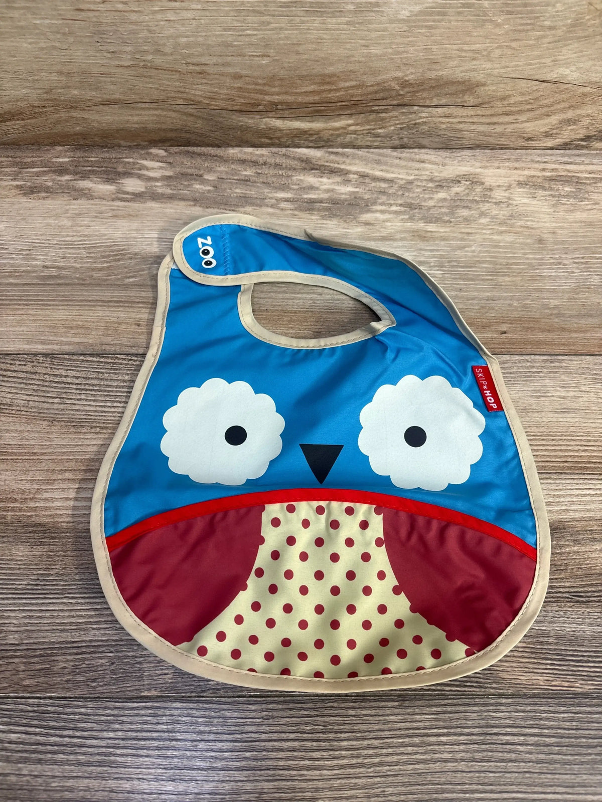 Flat lay of Skip Hop Zoo Tuck-Away Bib owl showcasing blue owl face, red polka-dot tuck-away pocket, beige trim on wood background