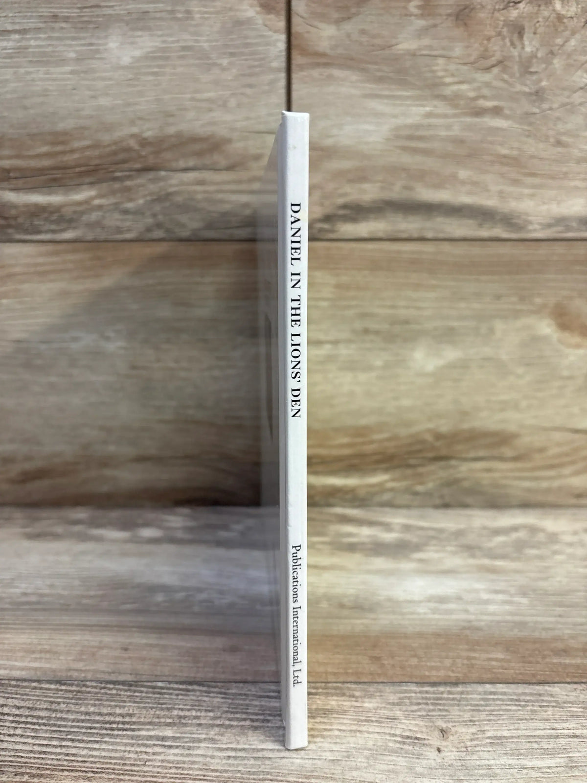 Spine view of Daniel in the Lions' Den hardcover children's book showing white spine with title and publisher on wood background