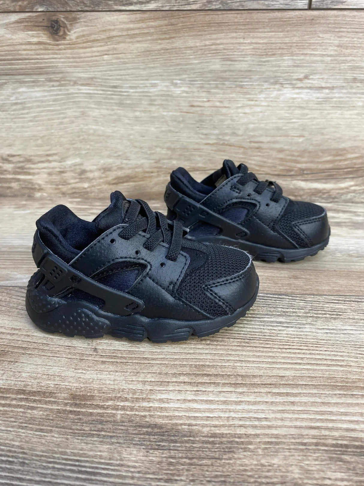 Front side view of toddler Nike Air Huarache Run triple black sneakers showing mesh and leather upper on wood background