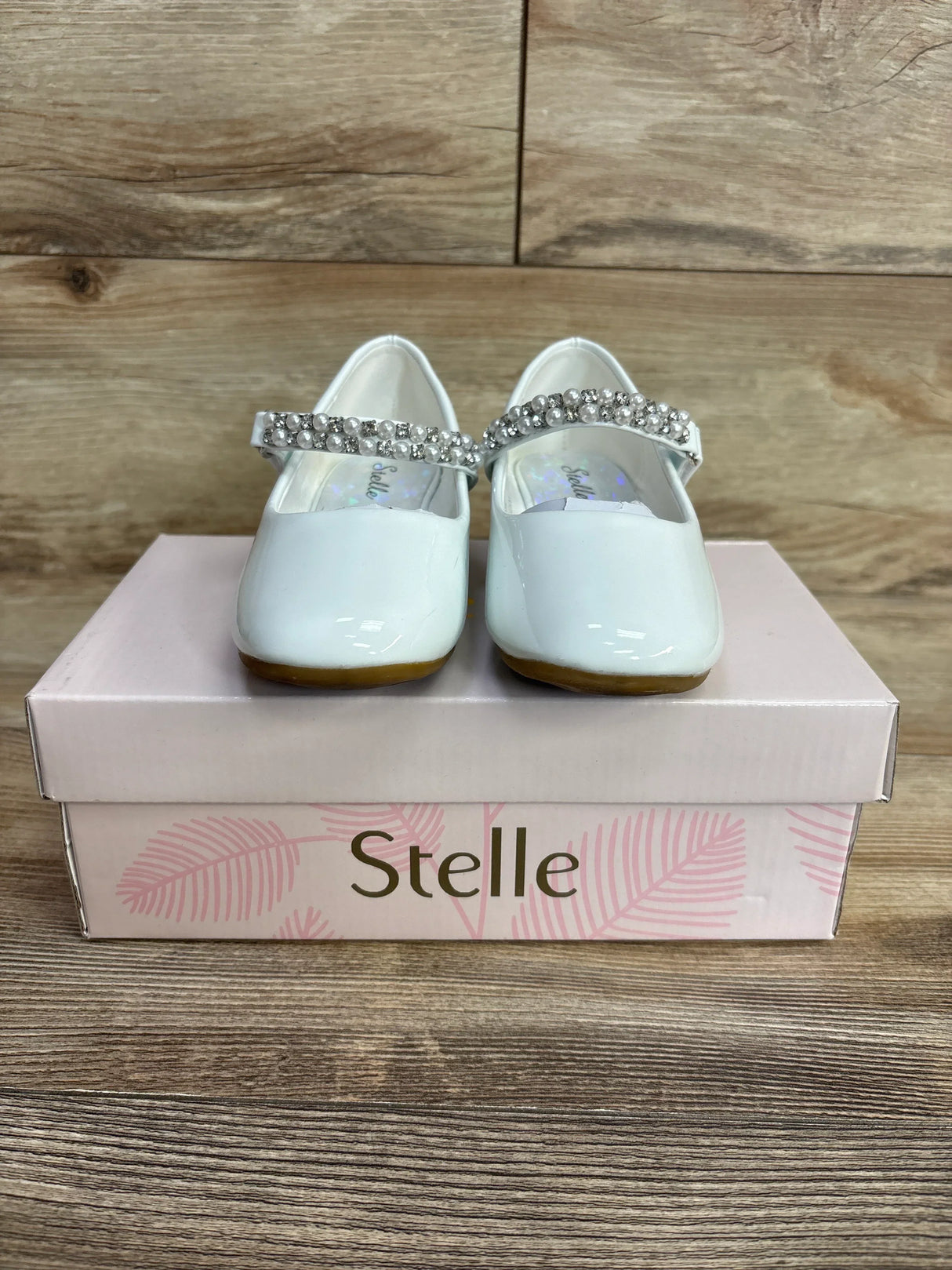 Pair of white patent leather toddler shoes with rhinestone straps on a pink Stelle shoebox