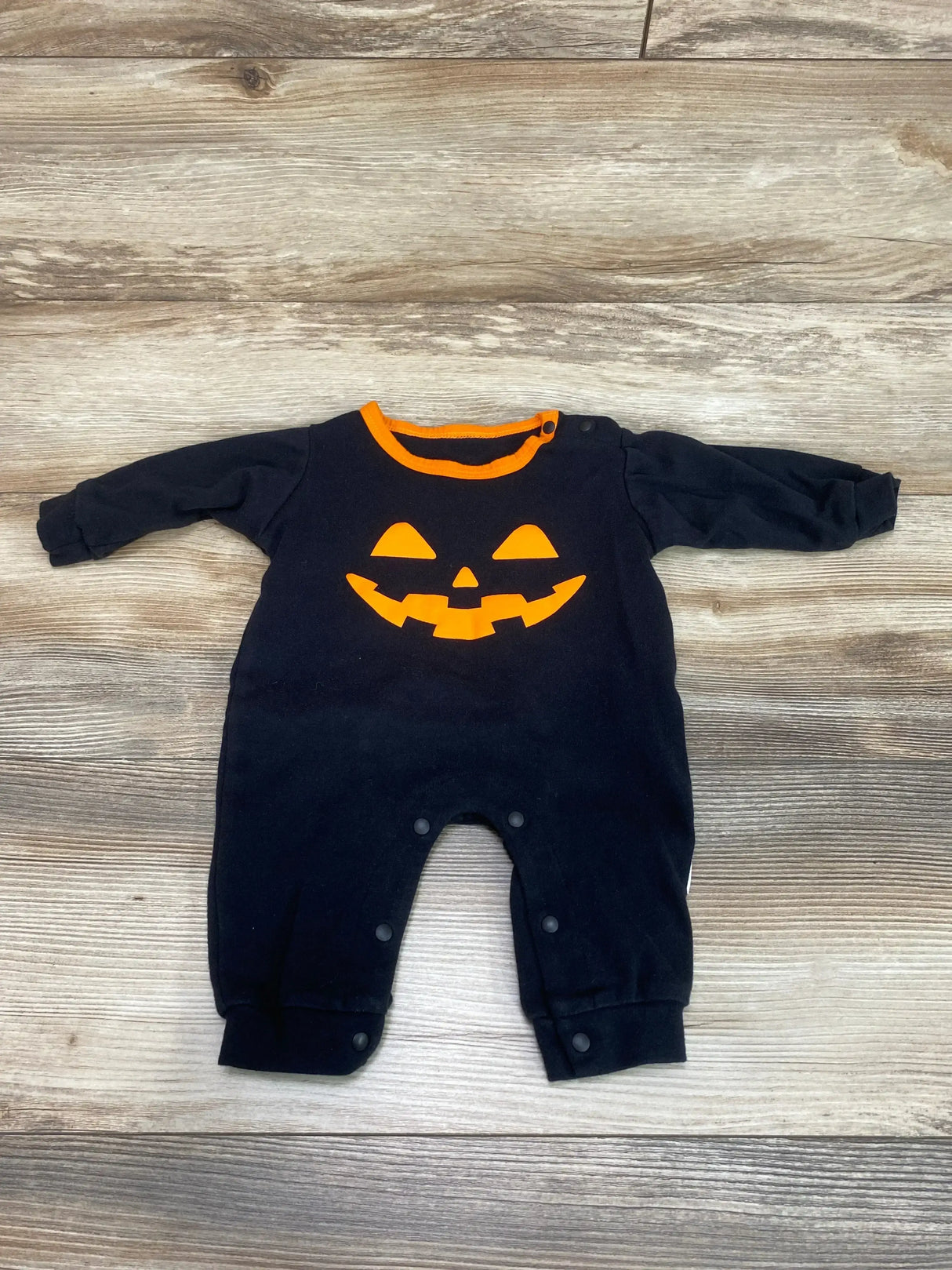 Flat lay of black baby coverall with orange jack o lantern face, orange neckline trim, and snap leg closures on wood background