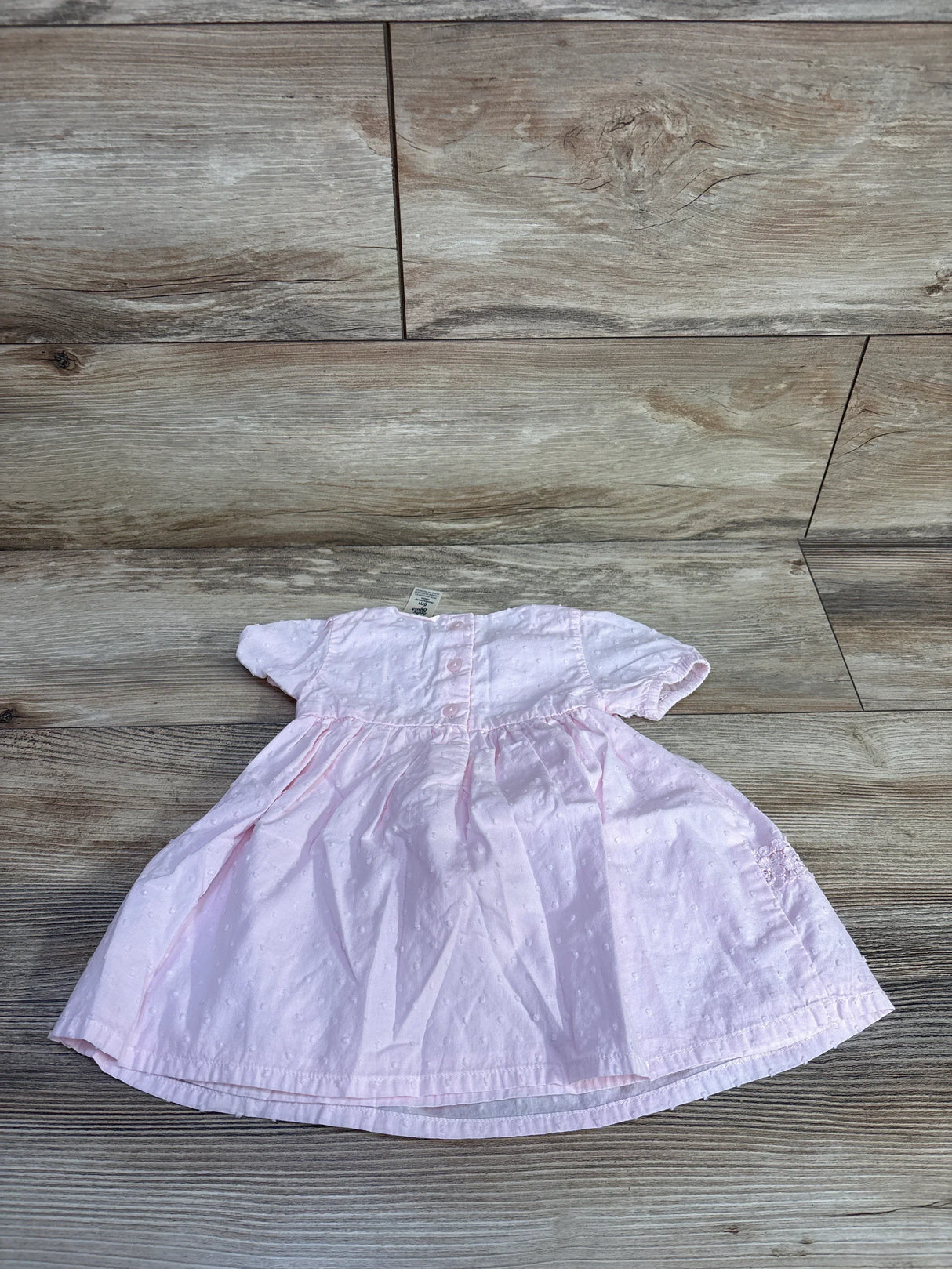 Flat lay of pink Oshkosh baby dress showing Swiss-dot texture, short sleeves, and back buttons on wood floor background