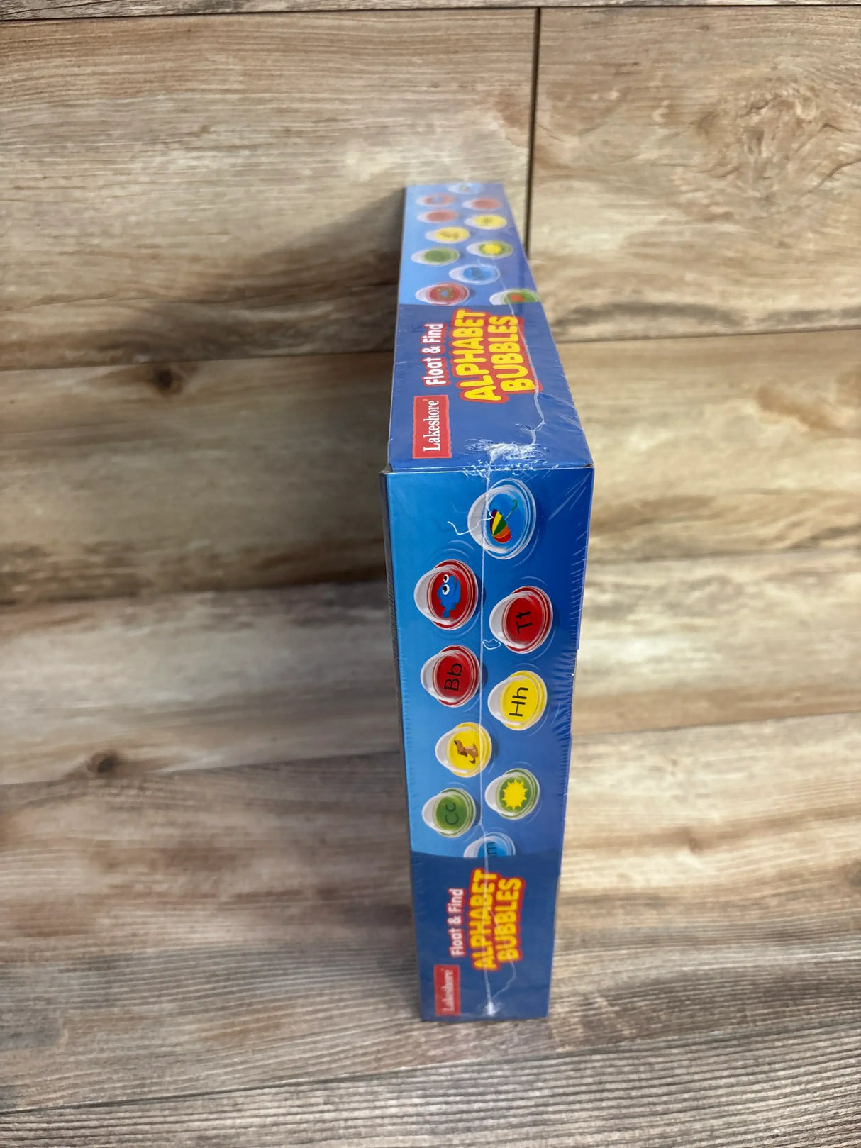 Side view of Lakeshore Float and Find Alphabet Bubbles box showing alphabet bubble icons and shrink-wrap tear on wooden tabletop