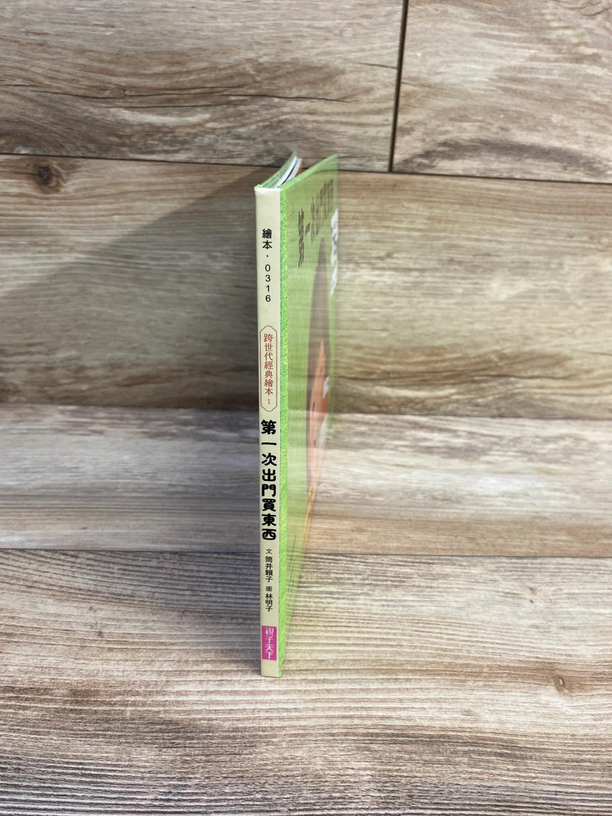 Spine view of Go Shopping for the First Time hardcover children’s book in Chinese showing thin green spine with Chinese title, standing on wood surface