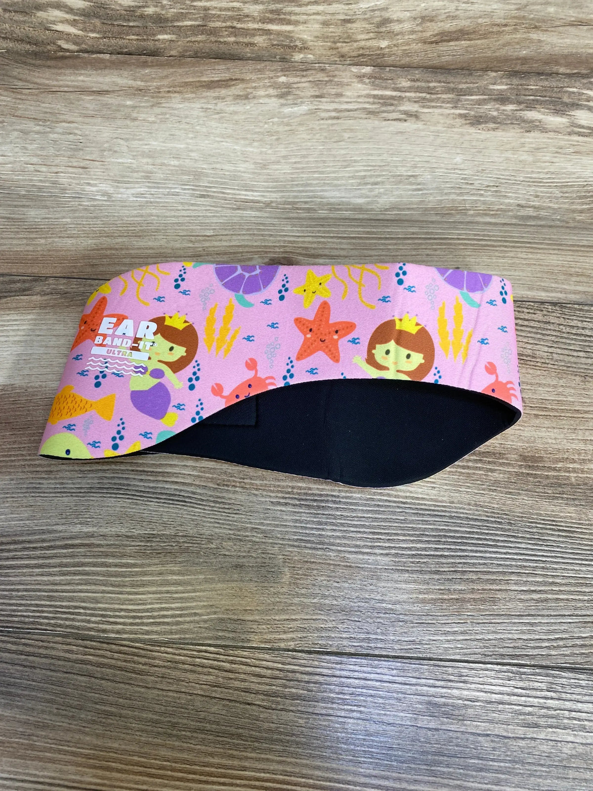 Flat-lay of pink Ear Band-It Ultra headband with mermaid and starfish print and black neoprene lining on wood background