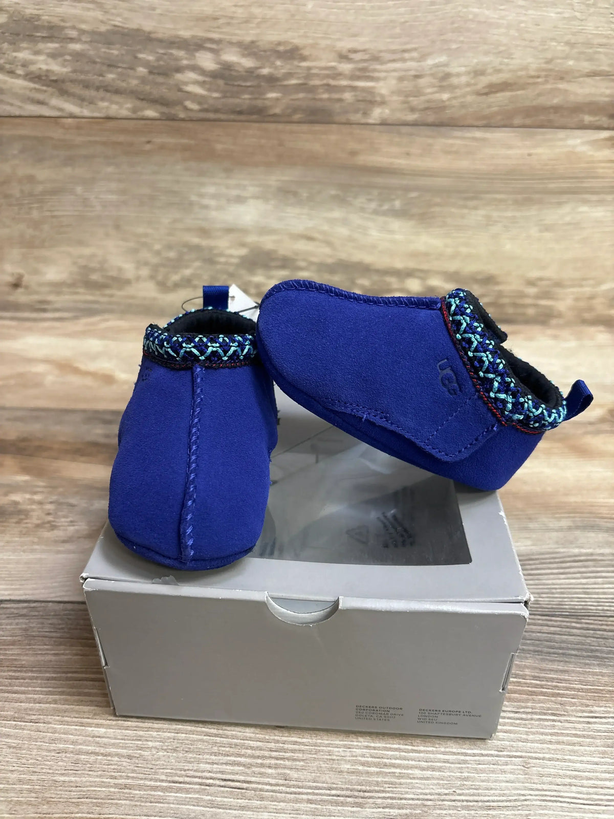 Side and front view of blue Ugg Baby Tasman slippers showing suede upper and patterned cuff on shoebox and wooden background