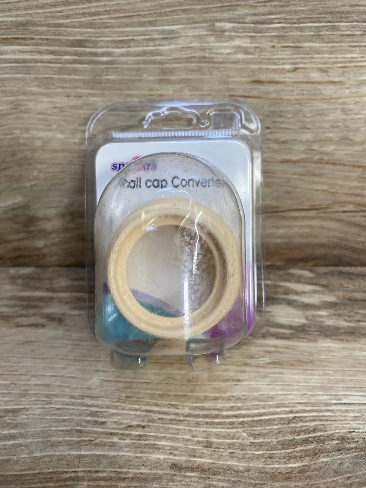 Top view of Spectra small-to-wide neck milk bottle cap adapter showing beige adapter ring in clear blister pack on wood background