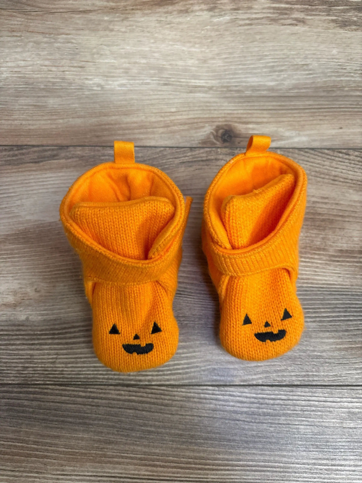 Top view of Cat & Jack Pumpkin crib booties size 3C showcasing orange knit fabric with black jack-o’-lantern faces on a wood floor