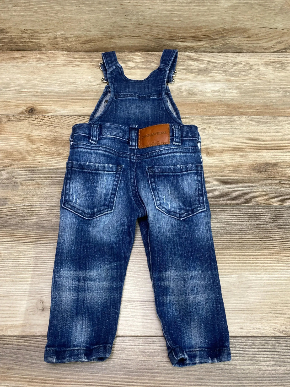 Back view of Dsquared2 baby denim overalls showcasing leather logo patch and rear pockets on wood plank background