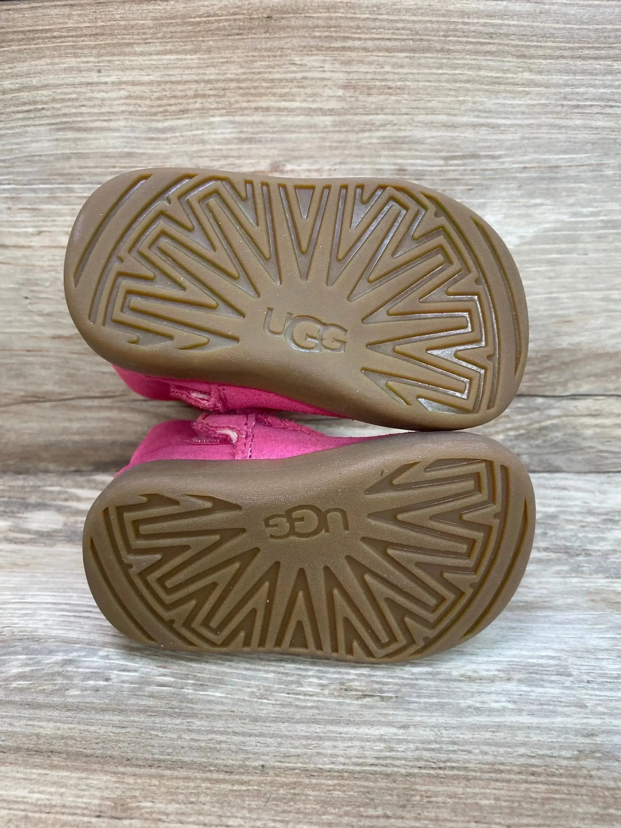 Bottom view of UGG Keelan toddler boots showing rubber tread pattern and UGG logo with pink suede uppers on wood background
