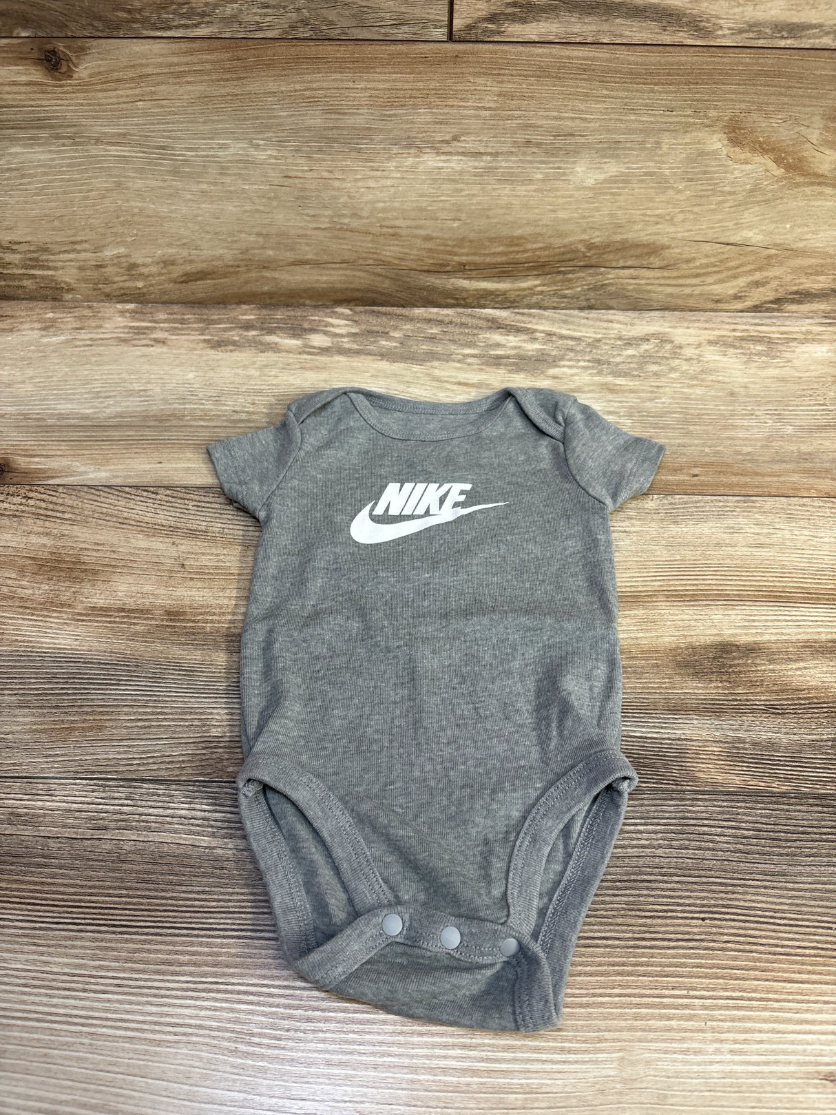 Top-down view of grey Nike bodysuit size 12 months showcasing white Nike logo and three snaps on wood background