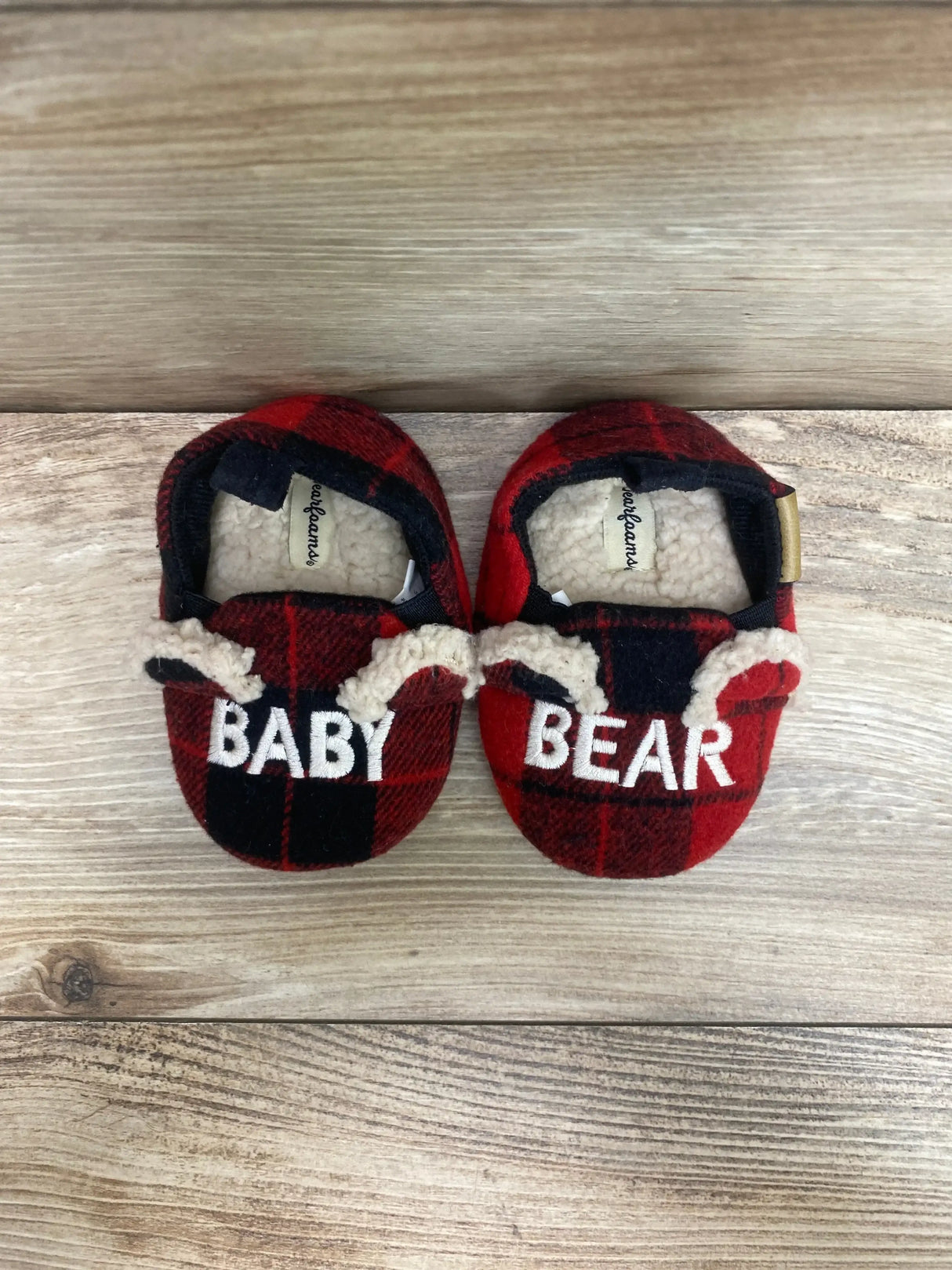 Top-down view of Dearfoams baby bear slippers, red and black plaid with embroidered BABY and BEAR and faux-shearling lining on wood background