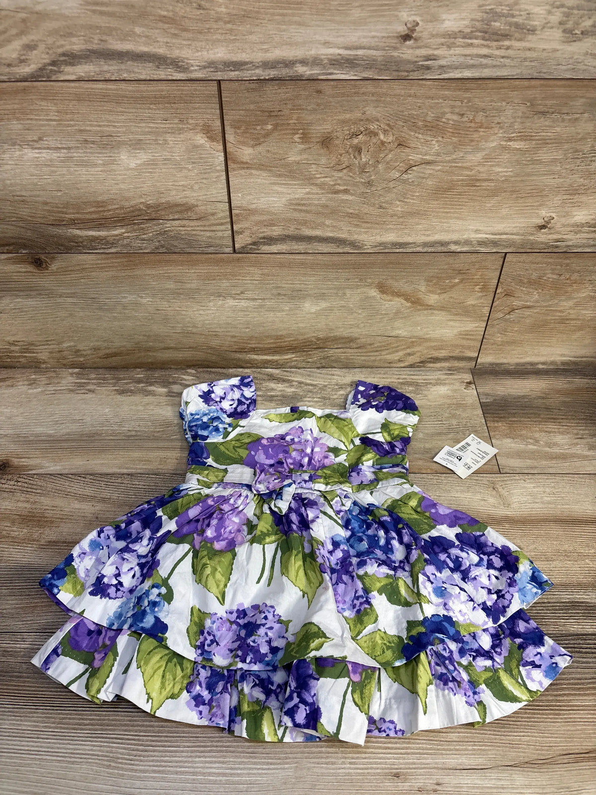 Front view of white Gymboree floral bow dress with flutter sleeves, shirred waist, bow belt, and ruffle hem on wood surface