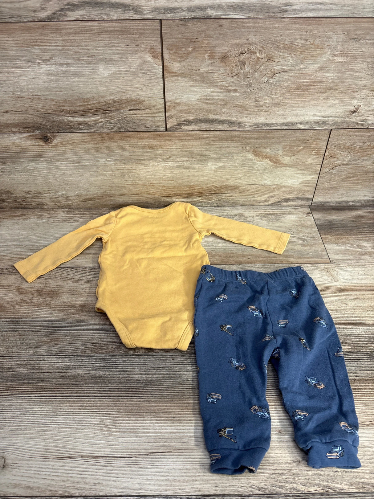 Yellow baby bodysuit with blue pants featuring construction vehicle print, laid on wood floor