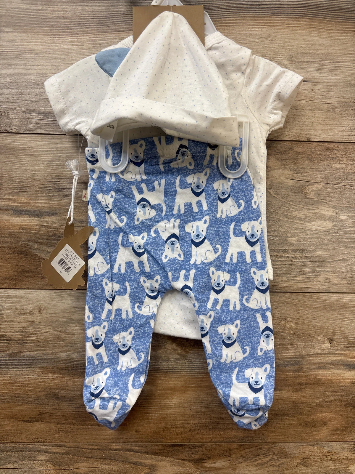 Top view of ED by Ellen DeGeneres three-piece dog layette set showcasing blue footed sleeper, dotted white hat and top on wood