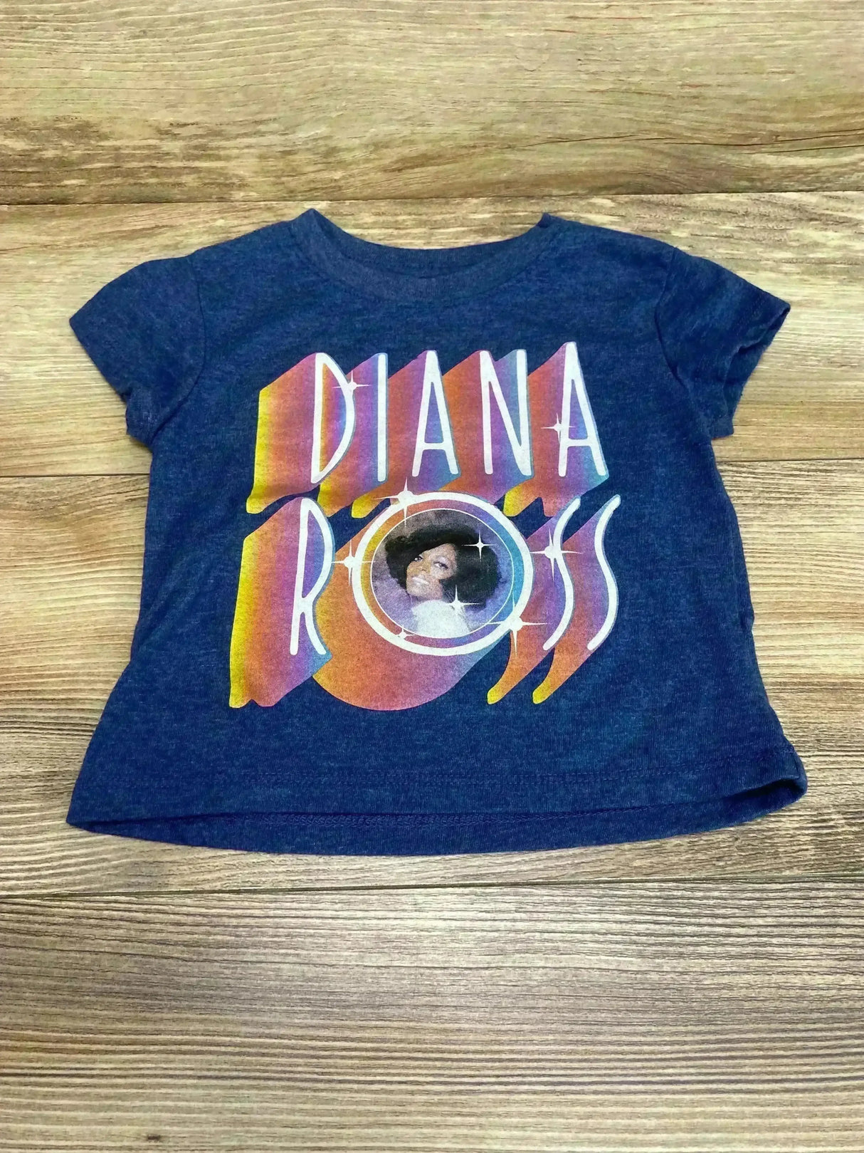 Front view of navy toddler Diana Ross t-shirt showcasing a large multicolor retro graphic with circular portrait, laid flat on wood background