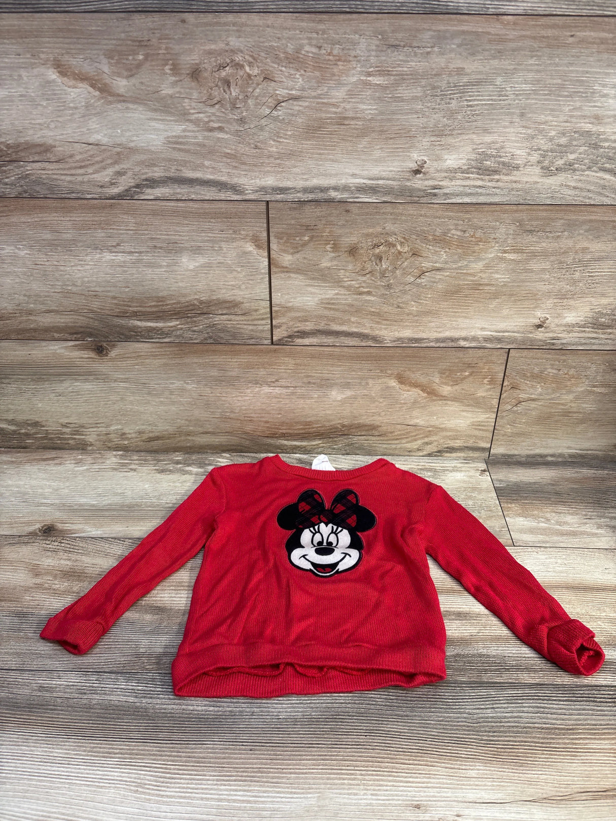 Front view of toddler red Minnie Mouse knit sweatshirt showing embroidered Minnie face with plaid bow, long sleeves, laid flat on wooden floor