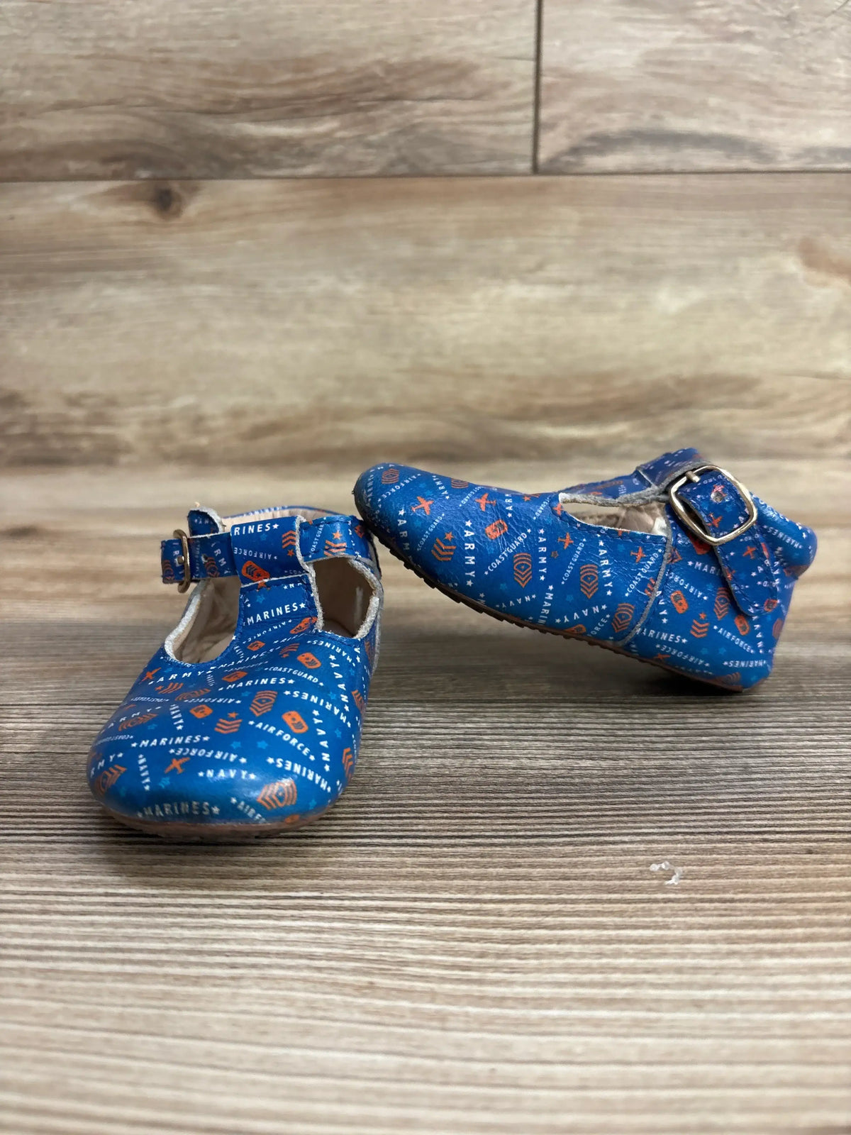 Three-quarter view of Monkey Feet T-Strap soft sole baby shoes showing blue military branch print and buckle on wood floor
