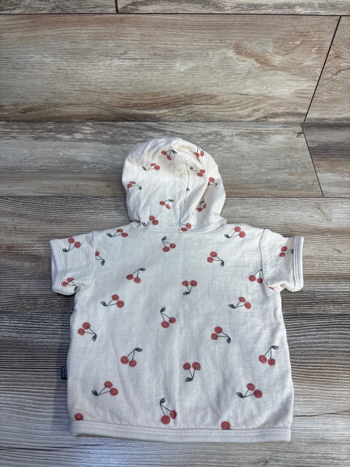 Back view of Modern Moments hooded shirt showcasing beige fabric, cherry print, short sleeves, and hood on a wood background