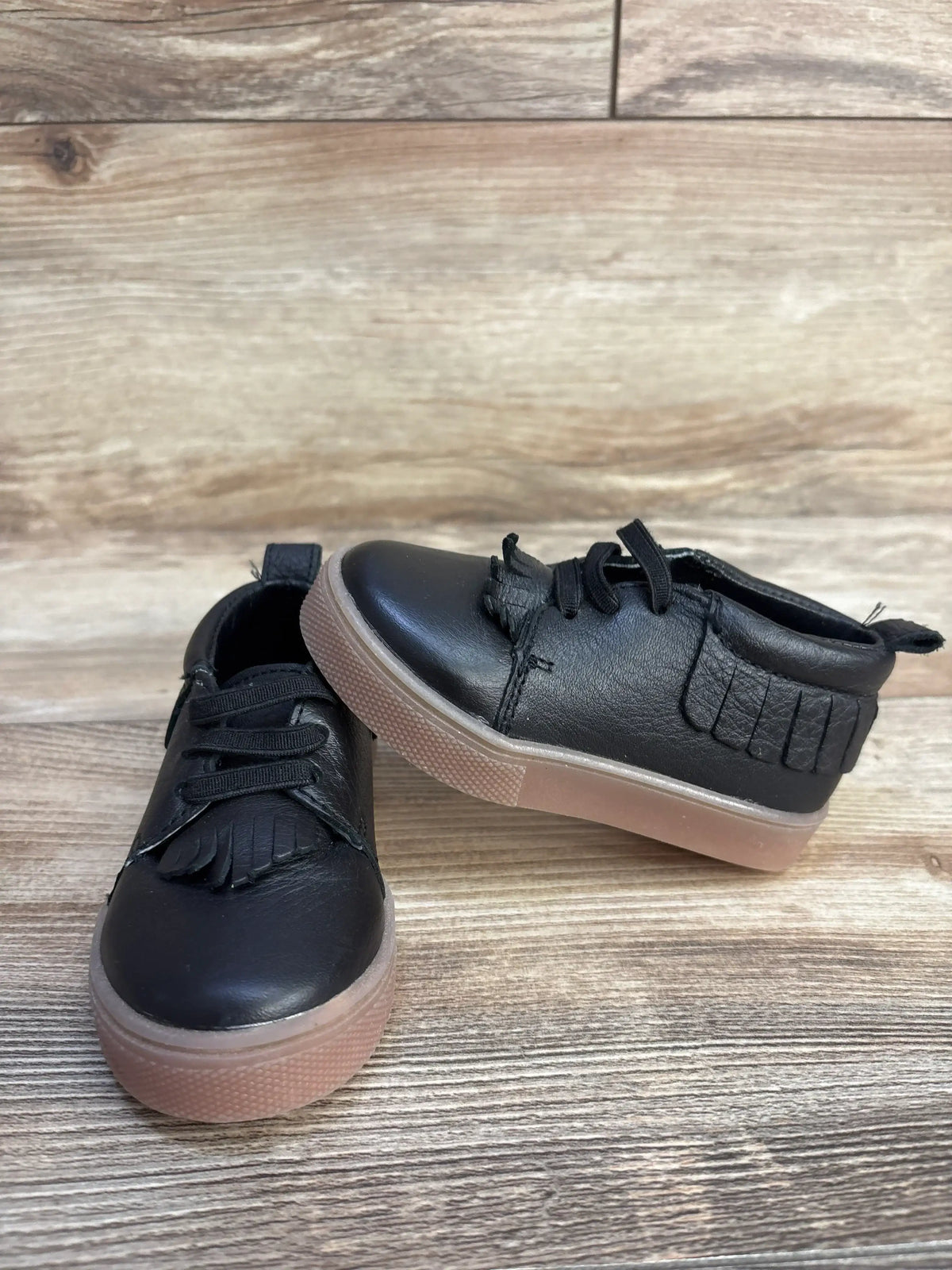 Front three-quarter view of Freshly Picked slip-on mid sneakers in black showing fringe kiltie and tan rubber sole on wood