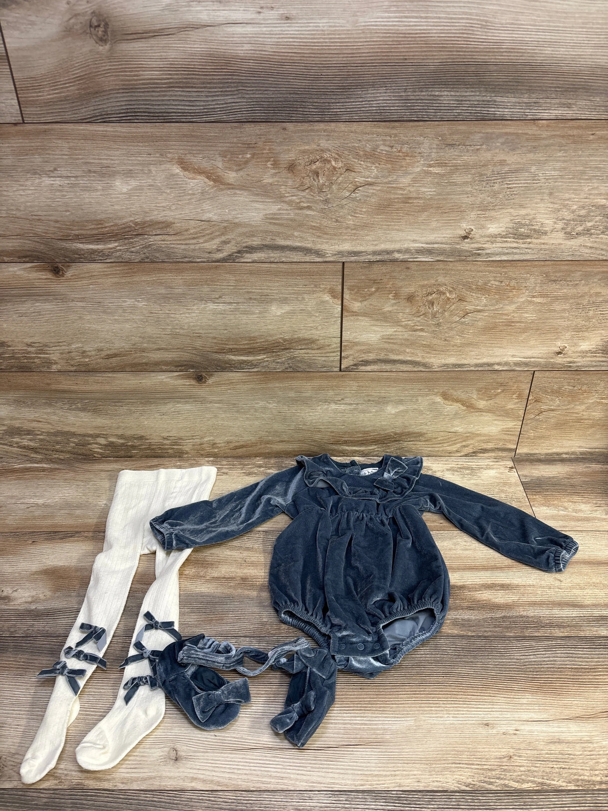 Flat lay of blue velvet romper set with ruffled collar, matching booties, and cream tights with bow accents on wood backdrop