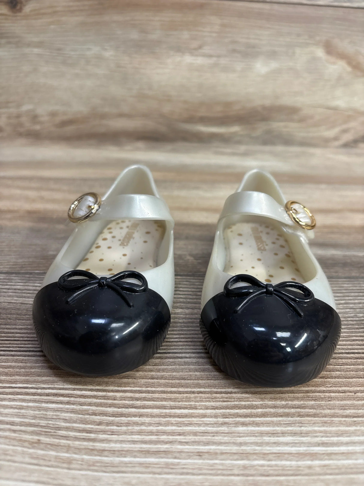Pair of cream and black toddler Mary Jane shoes with bows and gold buckle on wood floor
