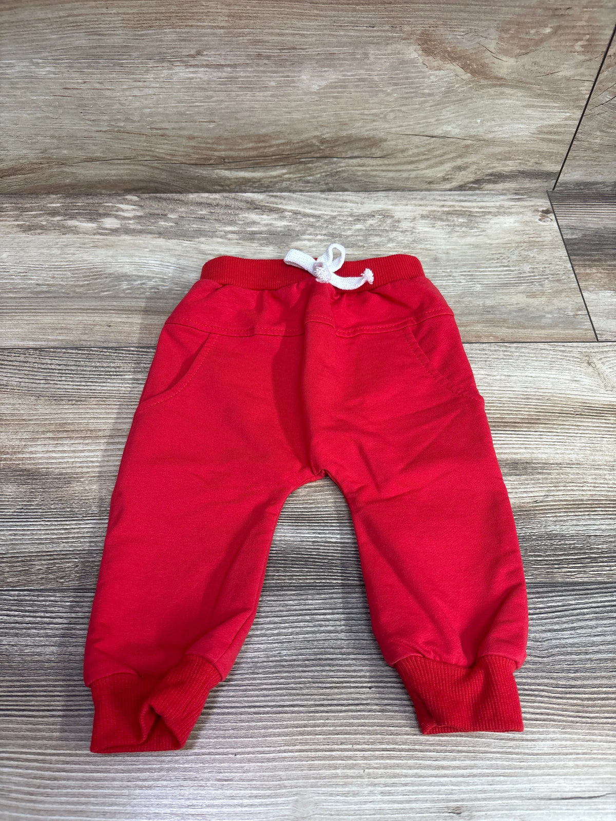 Front view of red drawstring harem pants for ages 3 to 6 months showcasing front pockets and ribbed cuffs on a wood-grain background
