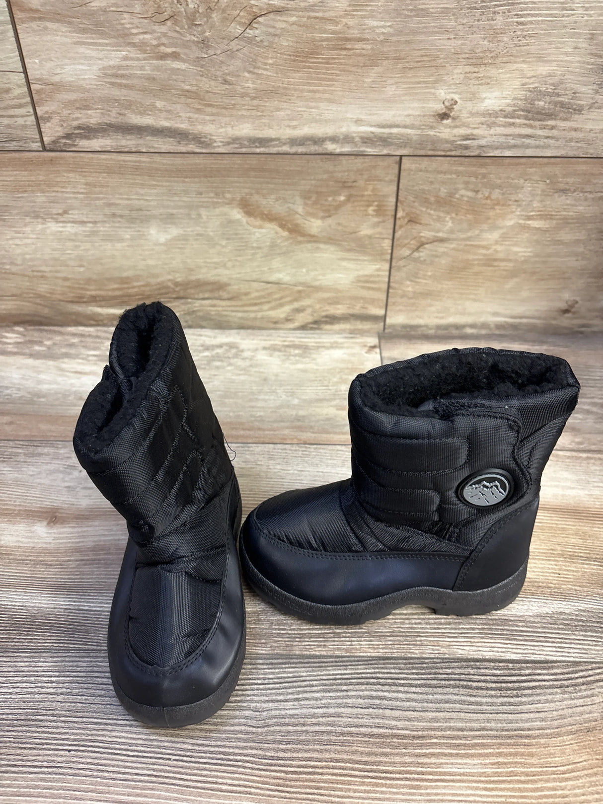 Three-quarter view of black toddler snow boots with quilted nylon shaft, faux-fur lining and rubber soles on wood floor