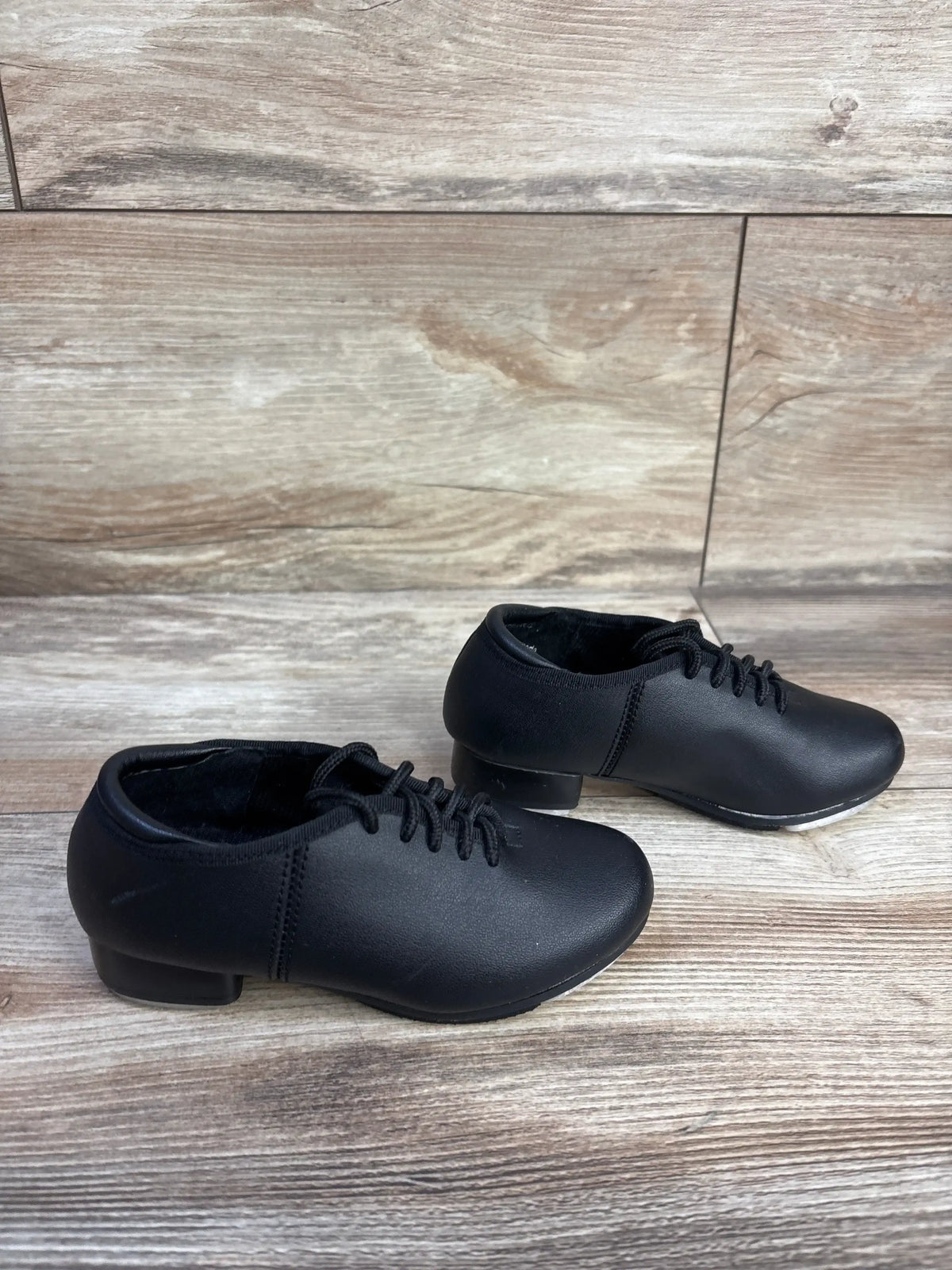 Side view of Theatricals kids black tap shoes, size 7C, showing laces, low heel and metal taps on a light wood background