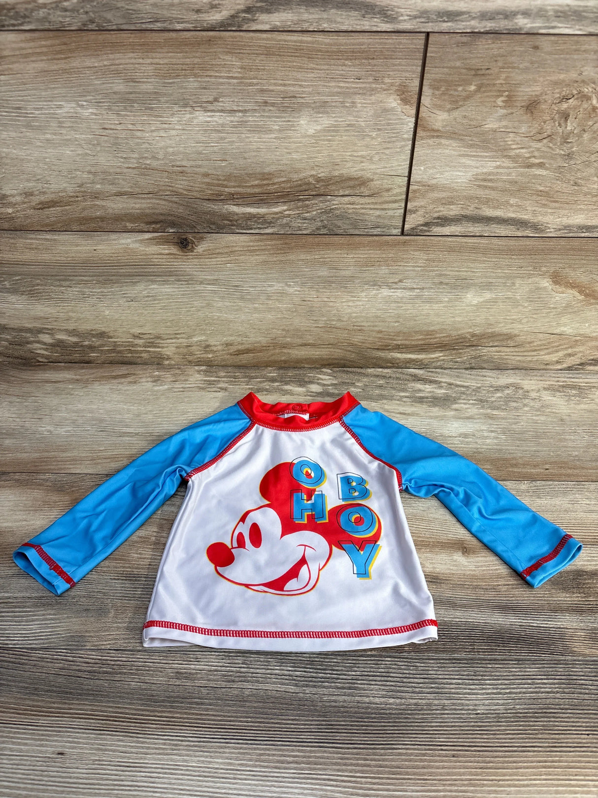 Front view of baby rashguard with large red Mickey face and OH BOY graphic, blue raglan sleeves and red trim on wood floor