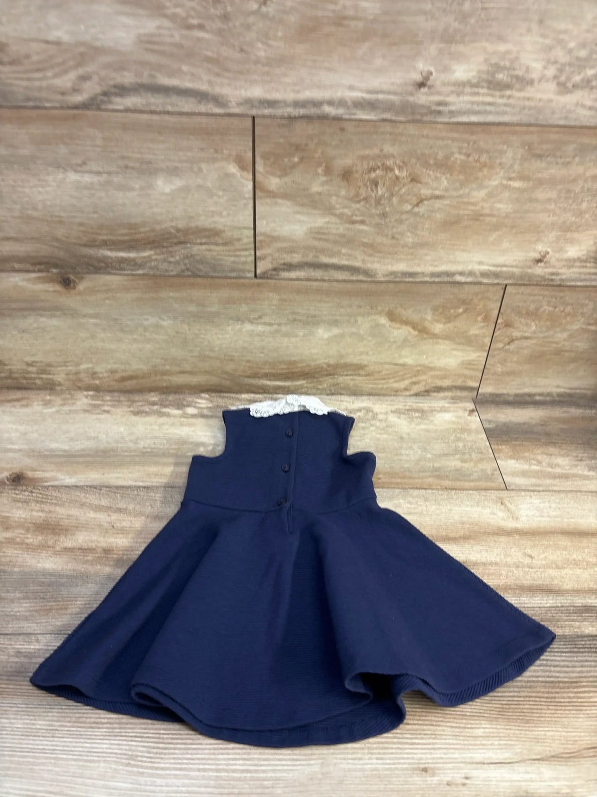 Front flat-lay of Janie and Jack navy sleeveless baby dress with white lace collar and flared skirt on wood background