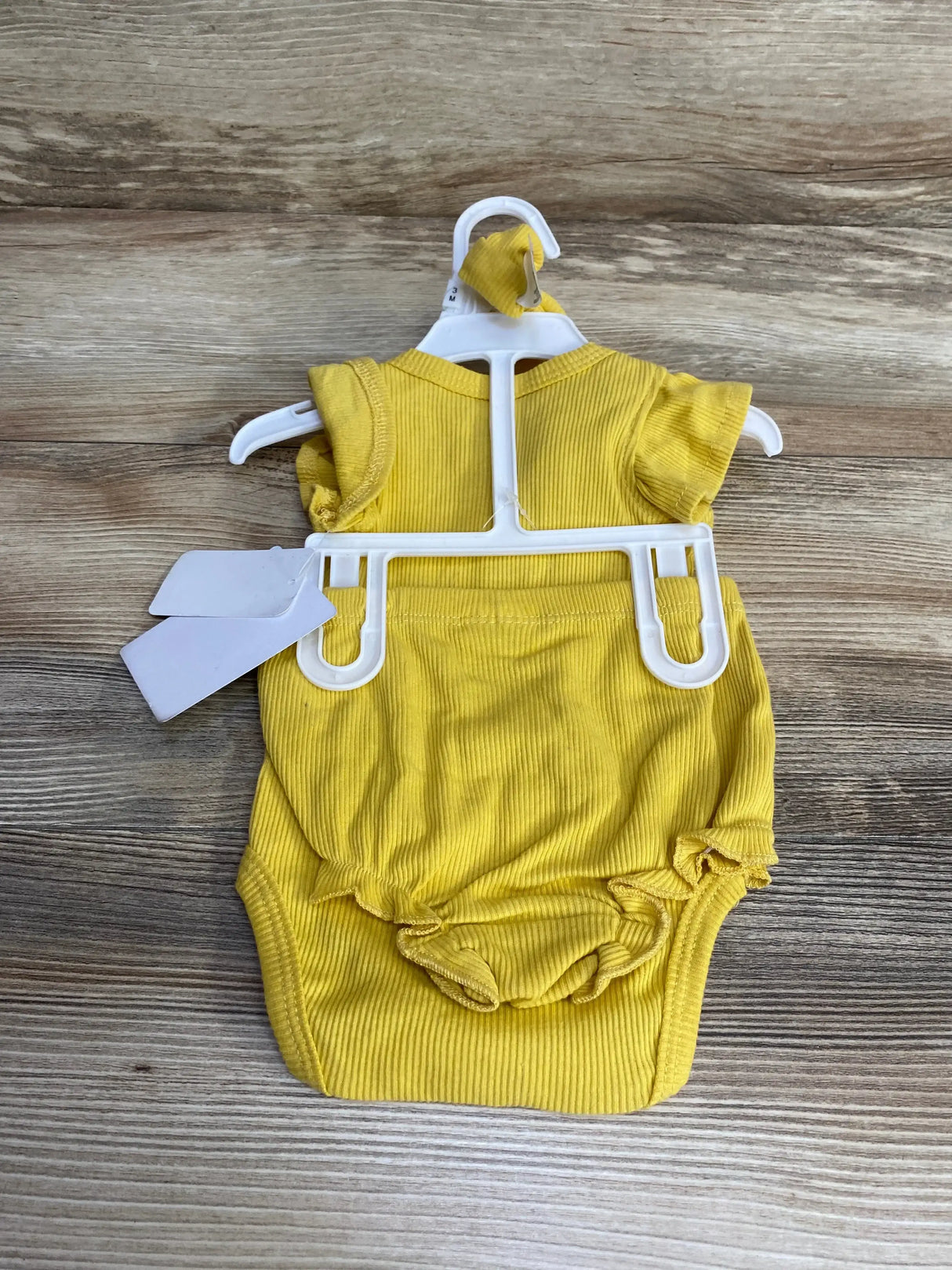 Top down view of yellow ribbed baby bodysuit size 3M on white hanger showing ruffle leg openings and short sleeves on wood background