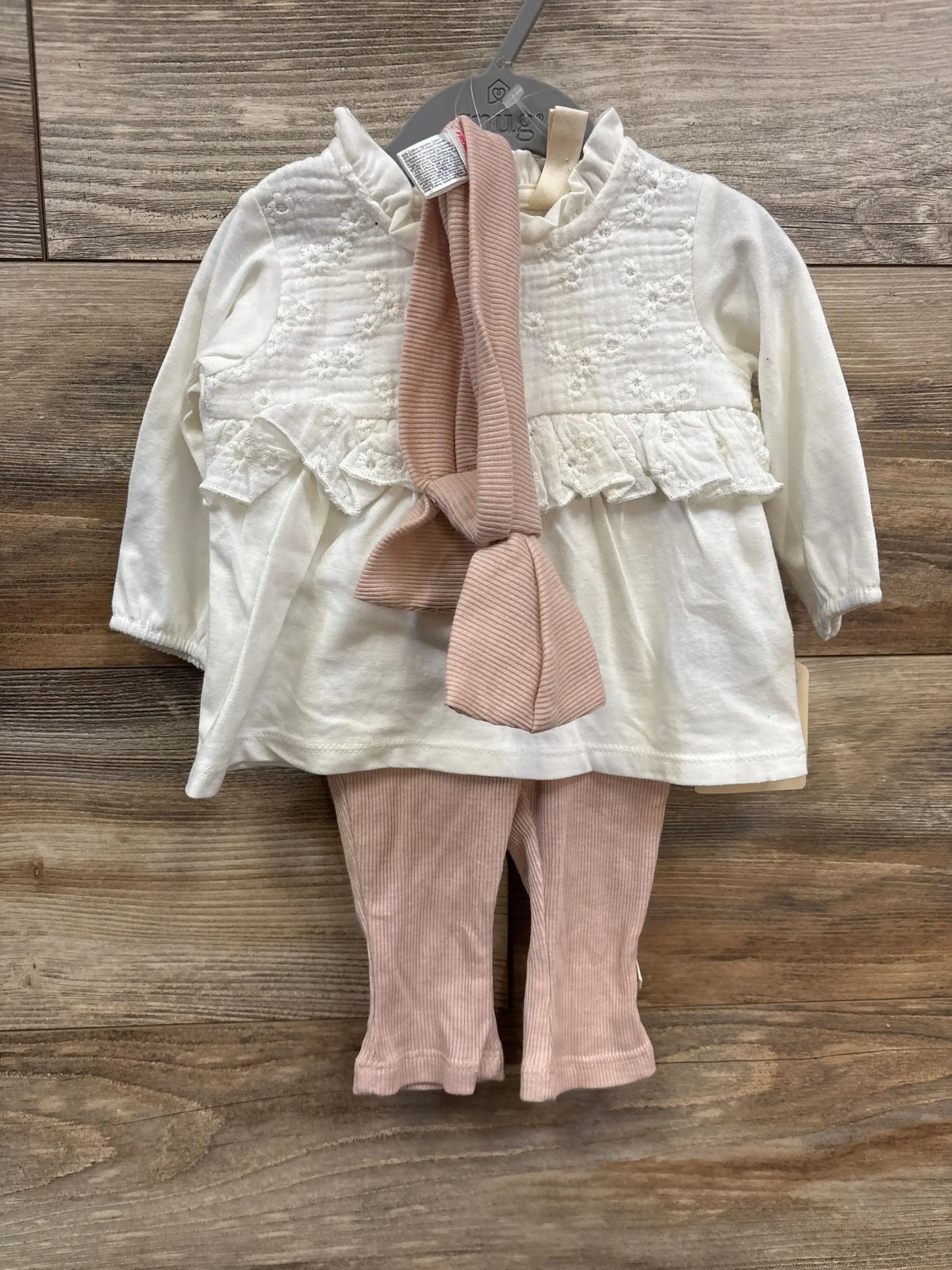 White long-sleeve baby top with floral embroidery and ruffles, paired with pink ribbed leggings and headband on wooden floor