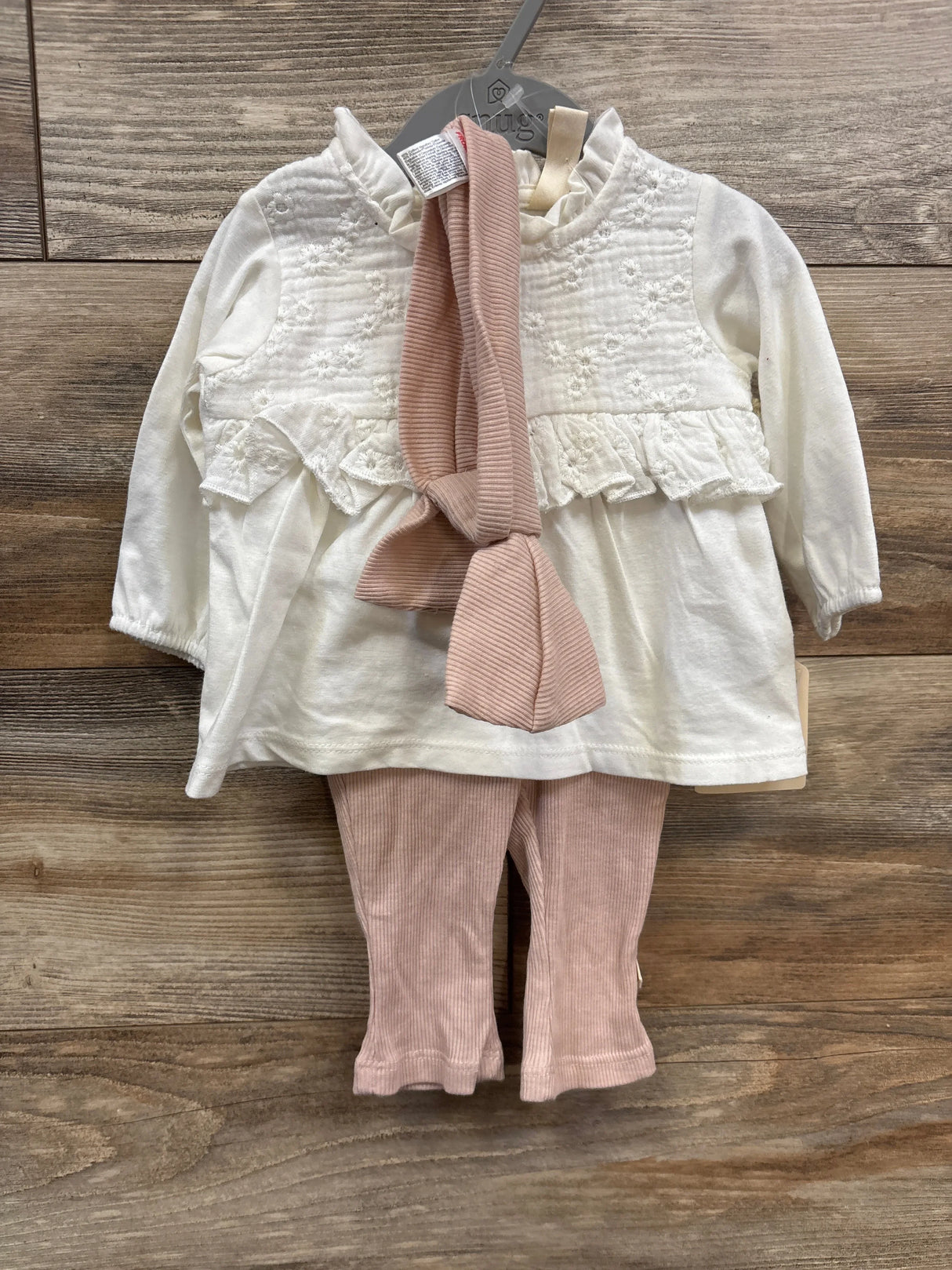 White long-sleeve baby top with floral embroidery and ruffles, paired with pink ribbed leggings and headband on wooden floor
