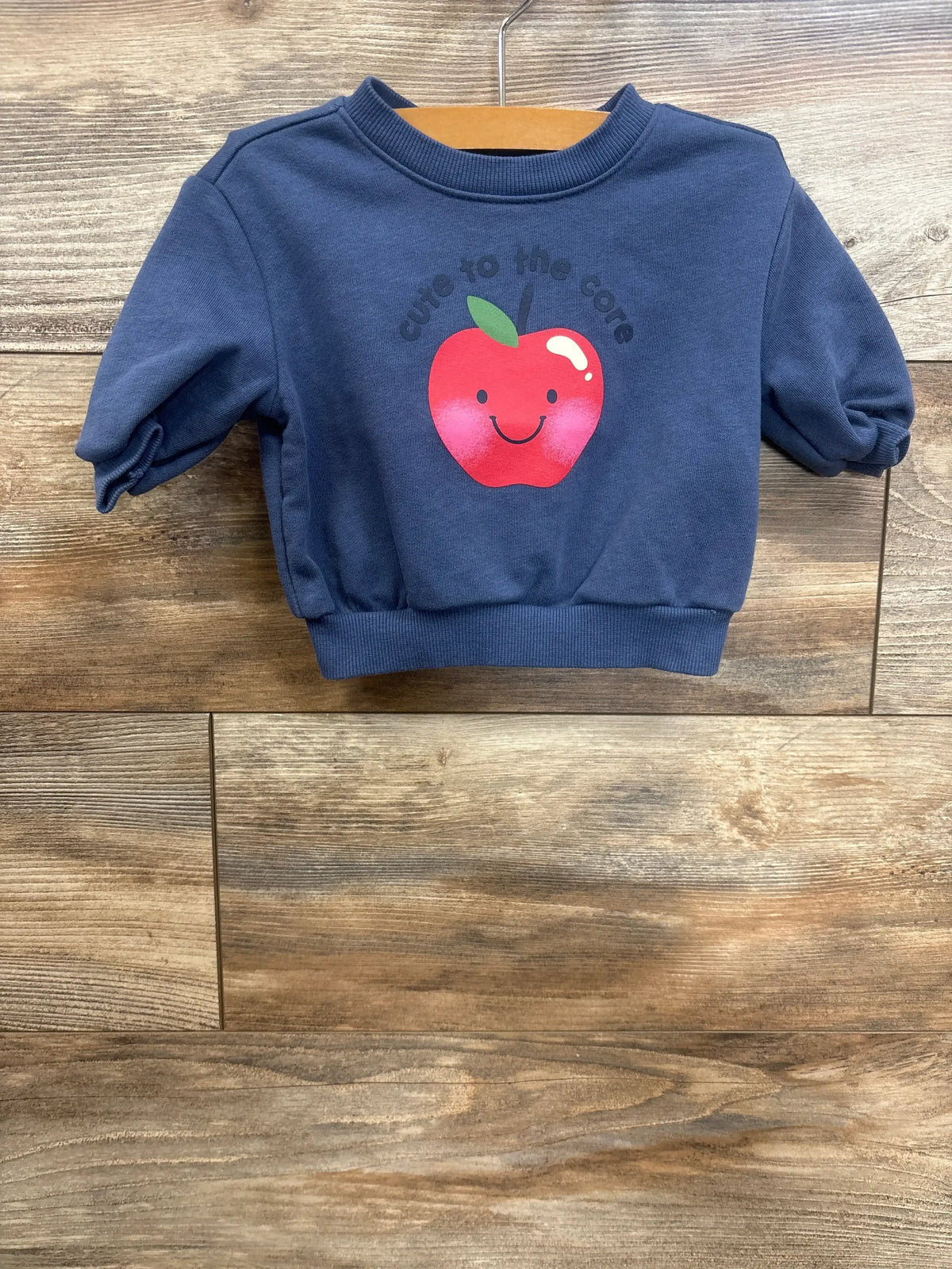 Front view of blue Cute To The Core sweatshirt, size 0-3m, showcasing apple graphic and ribbed cuffs on a wood background
