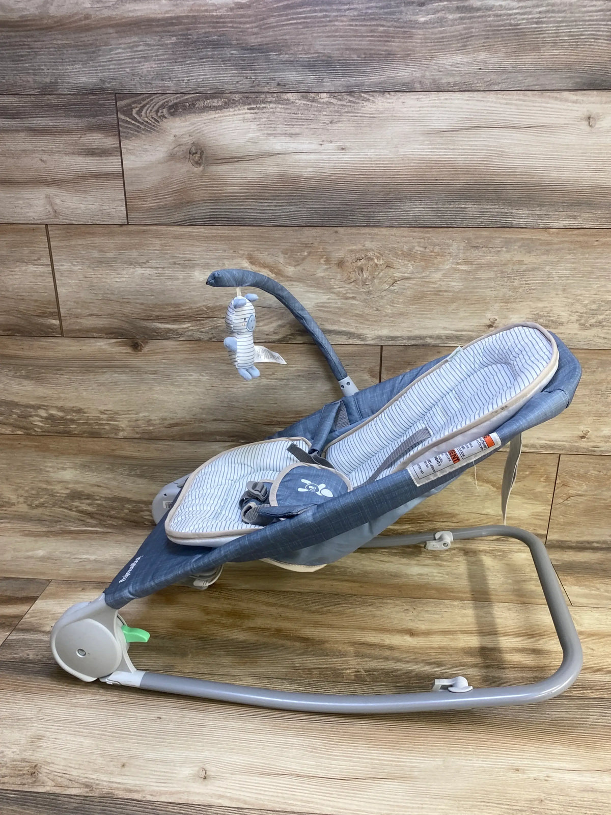 Side view of Ingenuity 2-in-1 Happy Belly bouncer showing blue-gray striped seat, toy bar with plush, and gray metal frame on wood background