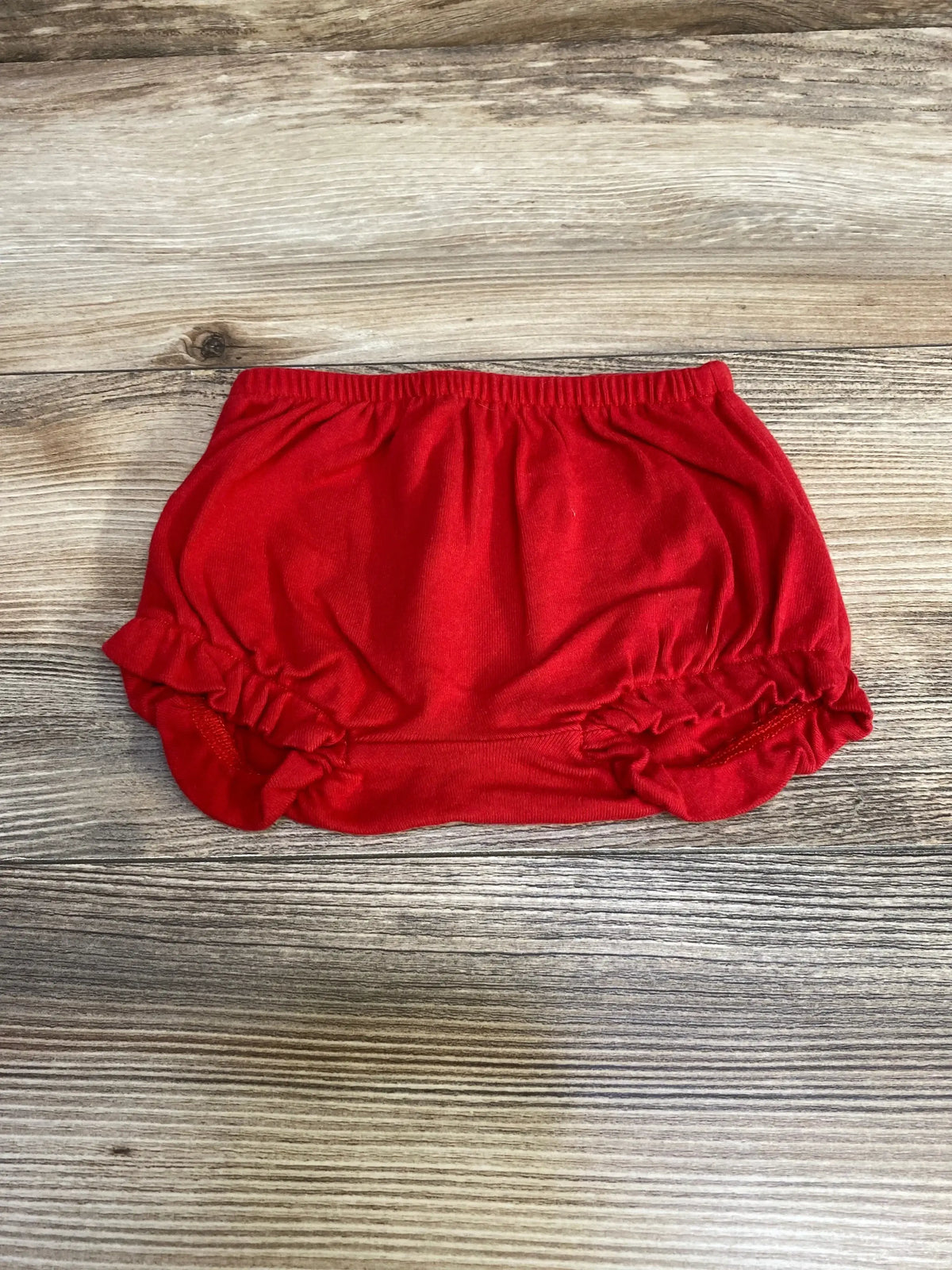 Top-down view of red baby bloomers size 3-6 months showing elastic waistband and ruffled leg openings on light wood