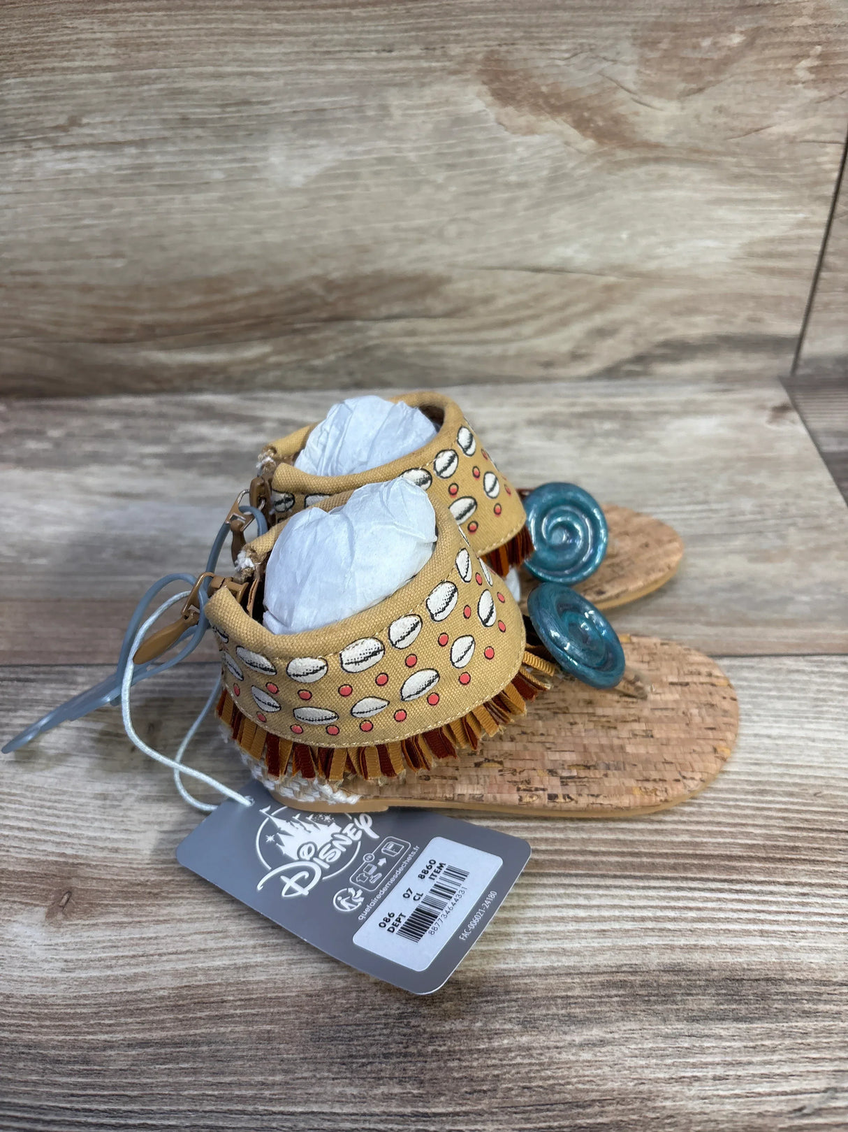 Three-quarter view of toddler Moana sandals with cork soles, tan shell-pattern ankle cuffs, brown fringe, and teal spiral toe accents on wood