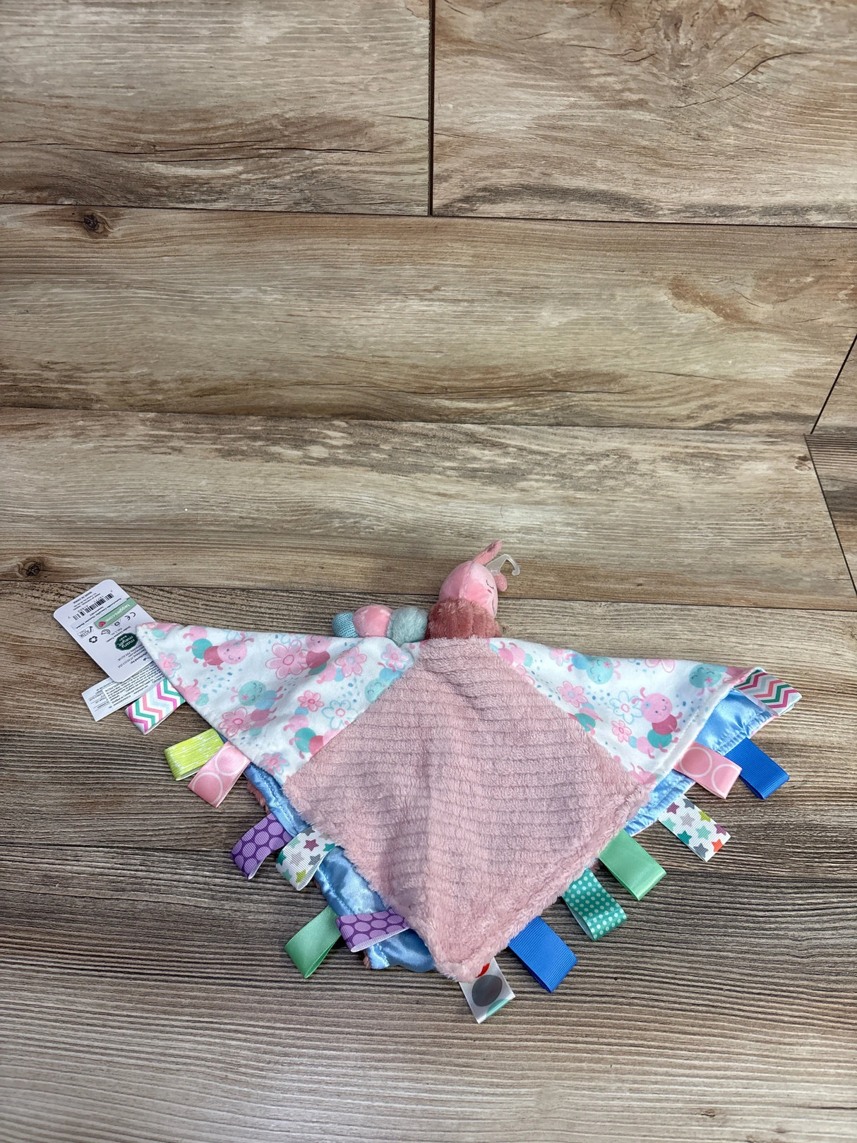 Soft pink and white baby sensory tag blanket with colorful ribbons and caterpillar head on wood floor