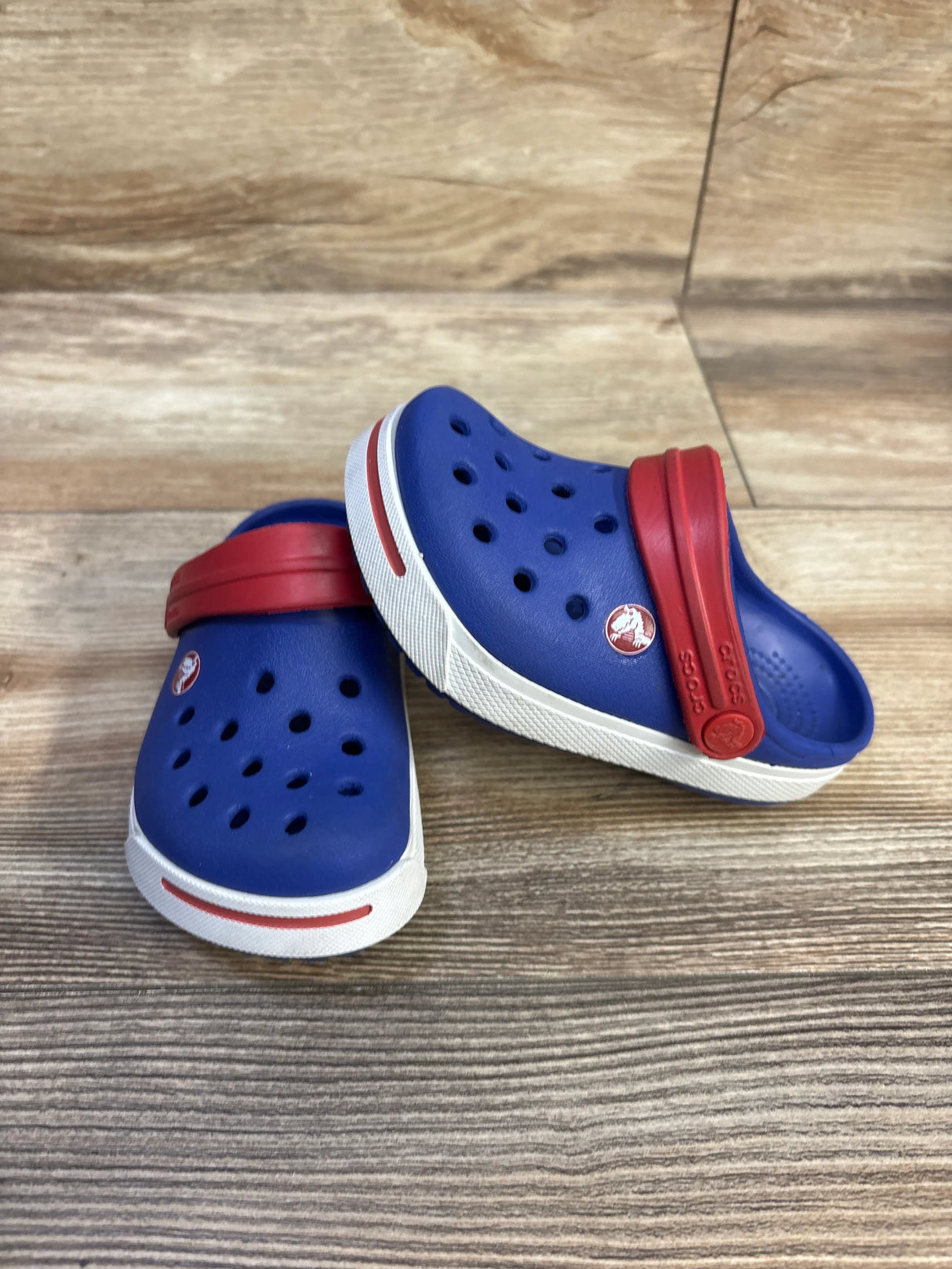Three-quarter view of blue Crocs Crocband II toddler clog size 6/7 with red heel strap and white sole on wood background