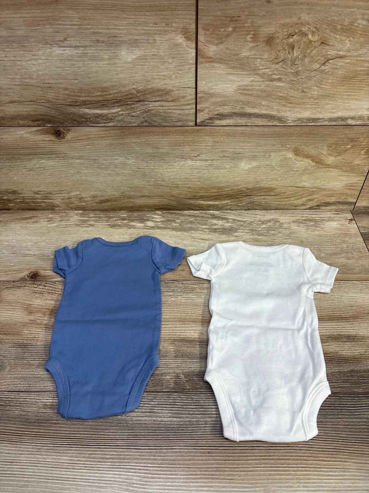 Front view of blue and white newborn bodysuits with short sleeves and bottom closures on a wood grain background