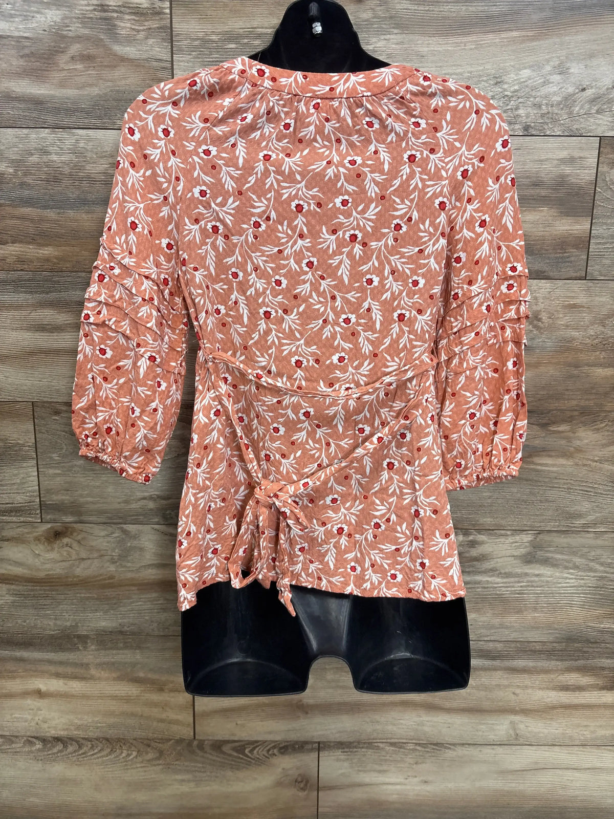 Back view of orange floral maternity blouse showing tie-back waist and gathered 3/4 sleeves on wood plank background