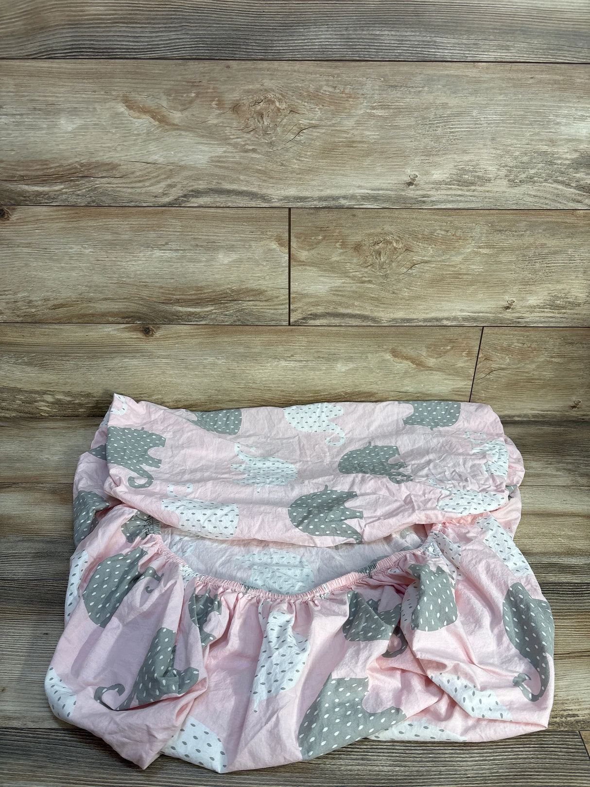 Top-down view of folded fitted crib sheet with pink fabric, gray and white elephant print and elastic edge on wood backdrop